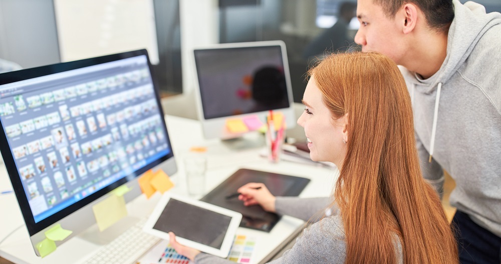 Zwei junge Webentwickler sitzen im Büro für Webdesign am Schreibtisch und besprechen die Gestaltung des neuen Onlineshops eines Kunden